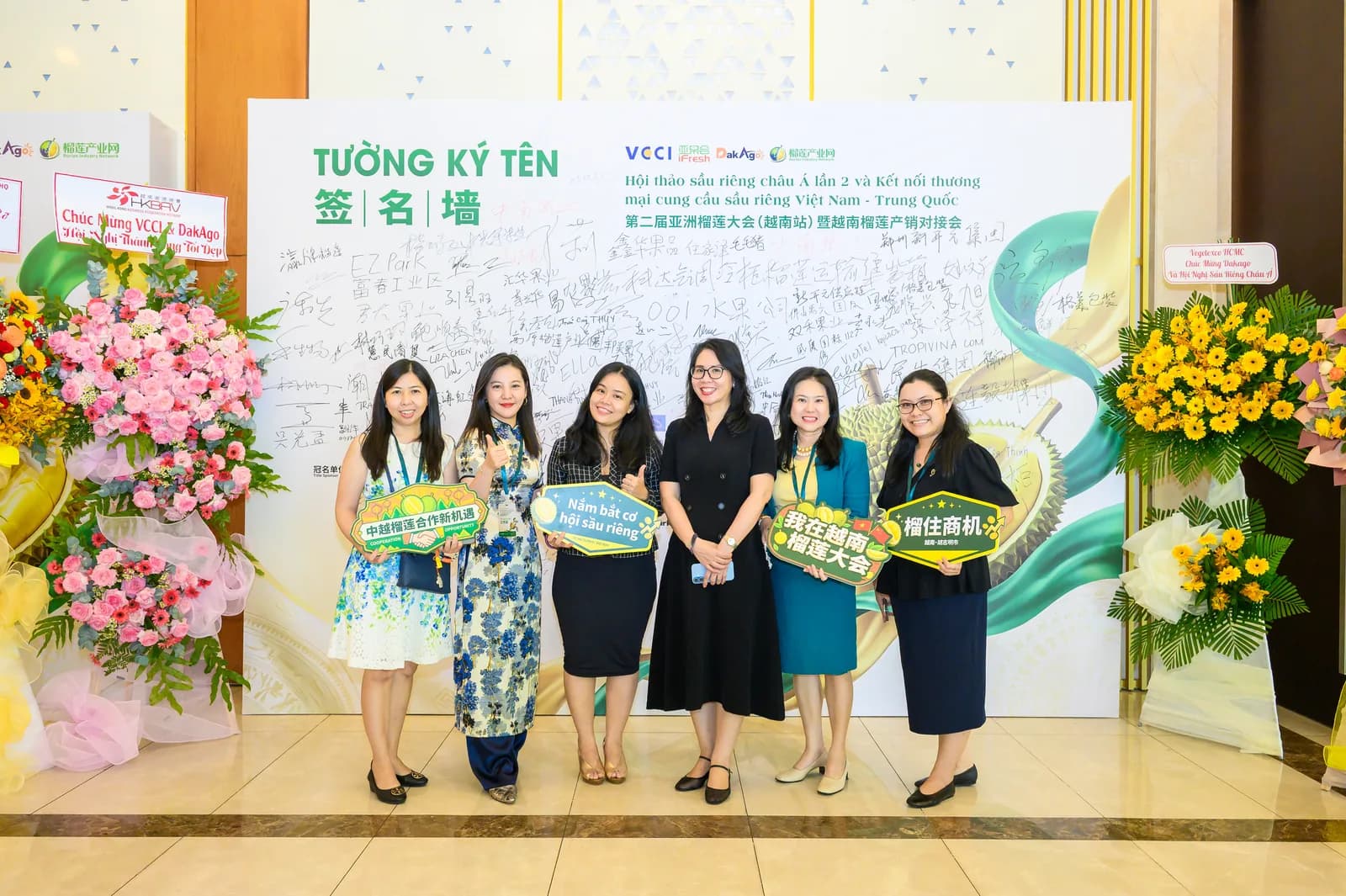 Nhóm nữ đại biểu chụp ảnh lưu niệm trước tường ký tên của Hội nghị, tay cầm các bảng chữ song ngữ Việt – Trung về cơ hội hợp tác sầu riêng
