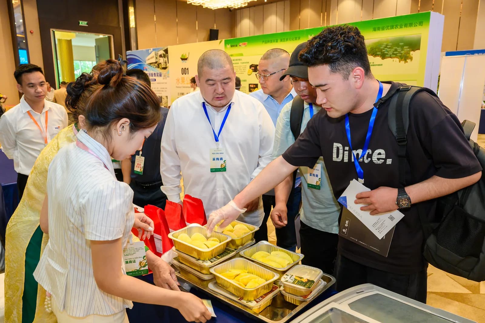 Các nhà thu mua Trung Quốc đeo thẻ đại biểu nếm thử múi sầu riêng vàng đựng trong khay tại khu trưng bày của Hội nghị Sầu riêng Châu Á lần thứ 2